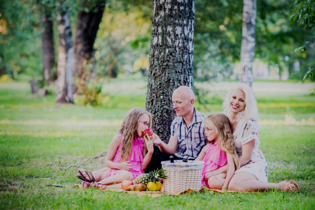 Perekond kahe lapsega peab pargis pikniku, kus nad on sättinud teki puu kõrvale ning istuvad teki peal.